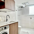 A modern bathroom interior featuring a walk-in shower with a frosted ribbed glass door, light gray stone tile walls and floor, a white toilet, a white washing machine, and a wooden vanity cabinet with a black faucet.