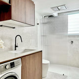 A modern bathroom interior featuring a walk-in shower with a frosted ribbed glass door, light gray stone tile walls and floor, a white toilet, a white washing machine, and a wooden vanity cabinet with a black faucet.