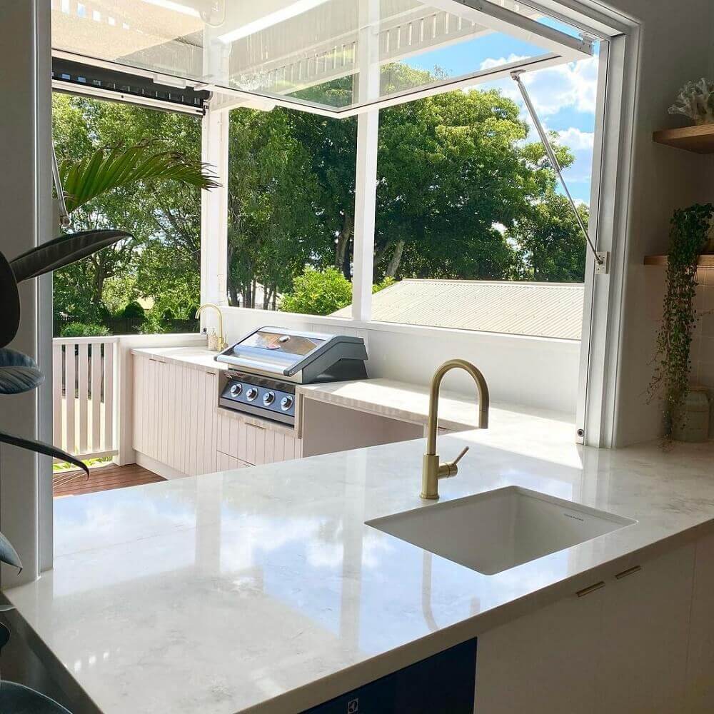 Modern kitchen with white marble countertop, sink, and grill area with large windows overlooking greenery.