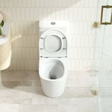 A modern white wall-mounted toilet with the seat lid open, set against a beige hexagonal tile wall and light stone floor. A glass shower door with gold hardware is visible on the right, and part of a vanity with flowers appears on the left.