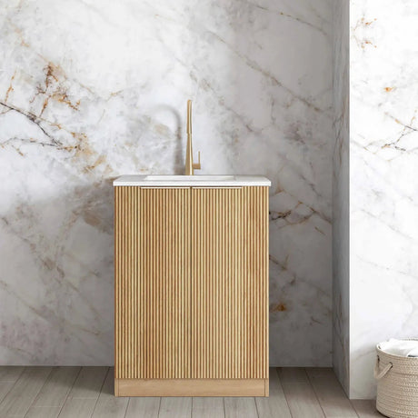 A modern bathroom vanity cabinet with a fluted light wood front, a white integrated sink top, and a tall matte gold faucet, set against a white marble wall with gold veining.