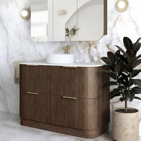 A modern bathroom vanity with a fluted brown wood cabinet, white countertop, white vessel sink, and a matching wall-mounted mirror. The vanity is set against a white marble wall and floor, with a potted plant on the right and round wall sconces on either side.