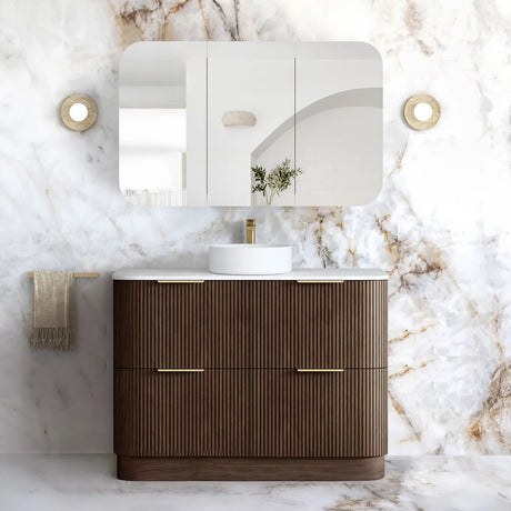 A modern bathroom vanity set featuring a dark brown ribbed cabinet with gold handles, a white countertop, a white vessel sink, a matching wall mirror, and a white marble-look wall and floor in a luxury bathroom setting.