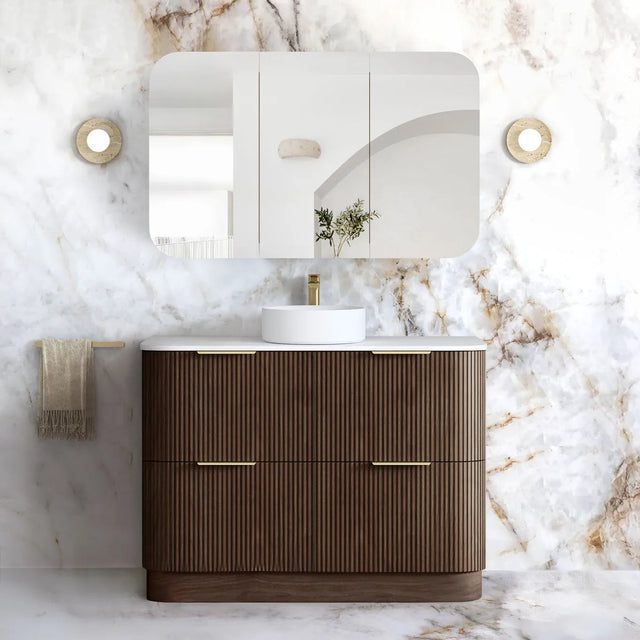 A modern bathroom vanity set featuring a dark brown ribbed cabinet with gold handles, a white countertop, a white vessel sink, a matching wall mirror, and a white marble-look wall and floor in a luxury bathroom setting.
