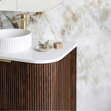 Modern bathroom vanity with a white vessel sink, gold faucet, fluted dark wood cabinet, white marble countertop, and a large oval mirror against a marble wall.