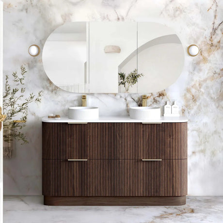 A modern double-sink bathroom vanity with a fluted dark wood cabinet, white countertop, and two white vessel sinks, set against a white marble wall with an oval mirror and wall sconces.