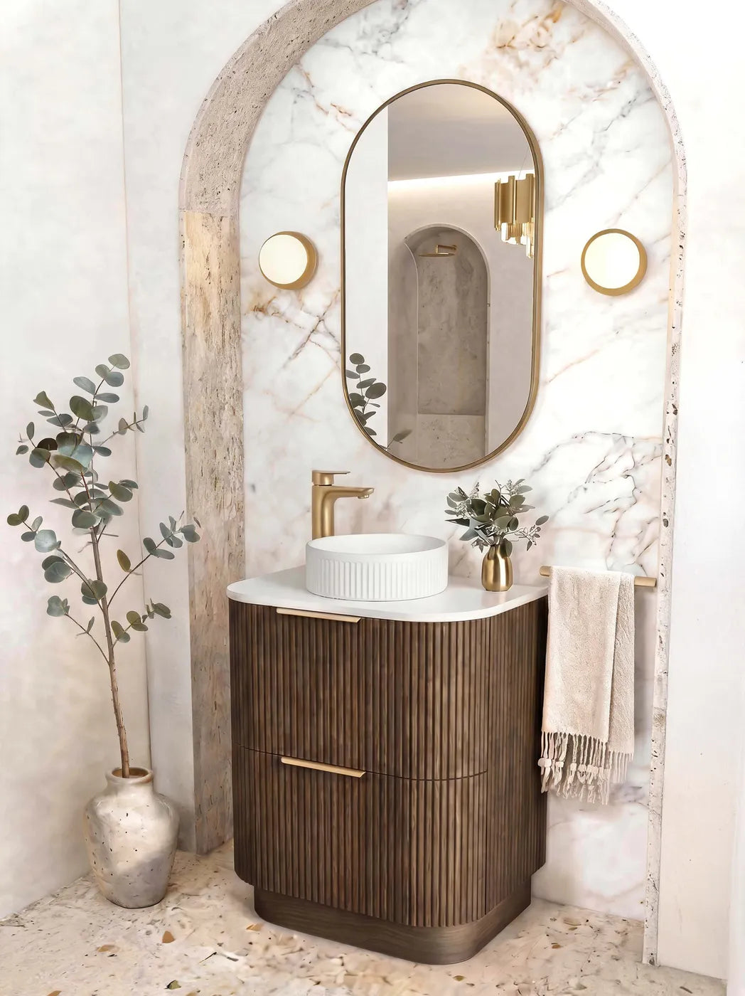 A modern bathroom vanity setup featuring a dark wood fluted cabinet with gold handles, a white vessel sink, a gold faucet, and an oval gold-framed mirror set within a marble-look arched niche.