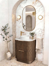 A modern bathroom vanity setup featuring a dark wood fluted cabinet with gold handles, a white vessel sink, a gold faucet, and an oval gold-framed mirror set within a marble-look arched niche.