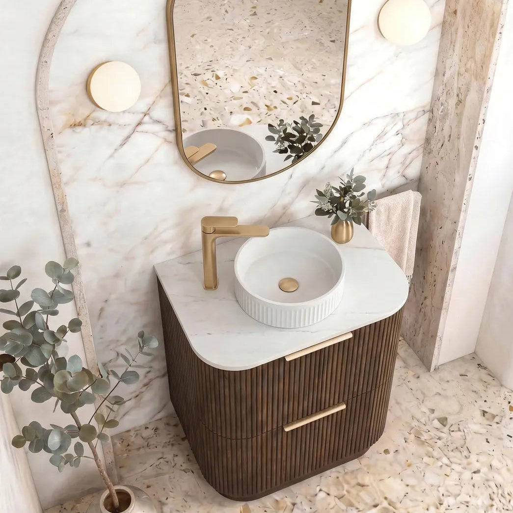 A modern bathroom vanity featuring a dark brown ribbed cabinet, a white marble countertop, a white round vessel sink, a gold faucet, and an oval mirror with a gold frame, set against a marble archway.