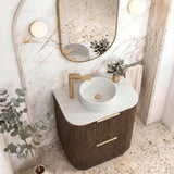 A modern bathroom vanity featuring a dark brown ribbed cabinet, a white marble countertop, a white round vessel sink, a gold faucet, and an oval mirror with a gold frame, set against a marble archway.