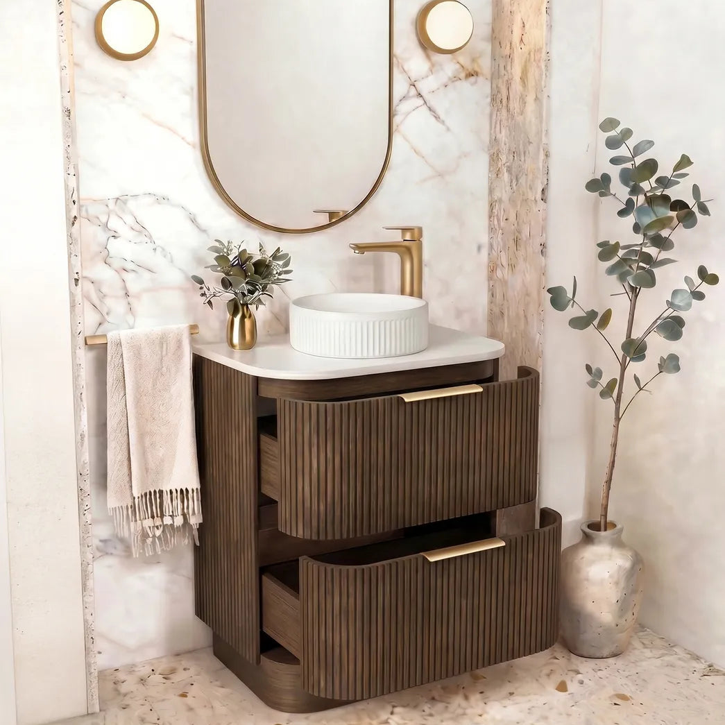 A modern bathroom vanity set featuring a dark brown ribbed wooden cabinet with gold-tone handles, a white ceramic vessel sink, a matching gold faucet, and an oval gold-framed mirror set against a white marble arched wall.