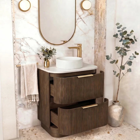 A modern bathroom vanity set featuring a dark brown ribbed wooden cabinet with gold-tone handles, a white ceramic vessel sink, a matching gold faucet, and an oval gold-framed mirror set against a white marble arched wall.