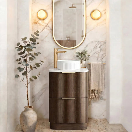 A modern bathroom vanity setup featuring a dark brown ribbed cabinet, a white vessel sink, a gold faucet, and an oval gold-framed mirror set against a white marble arch with wall sconces.