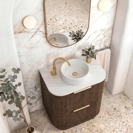 A modern bathroom vanity setup with a round white ceramic sink, a curved dark brown ribbed cabinet, a gold faucet, and an oval gold-framed mirror set against a marble-patterned wall.