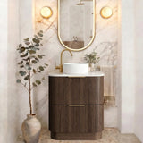 A modern bathroom vanity setup featuring a dark brown fluted cabinet, a white vessel sink, a gold faucet, an oval gold-framed mirror, and warm wall sconces against a marble-look arched backdrop.
