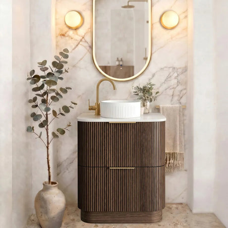 A modern bathroom vanity setup featuring a dark brown fluted cabinet, a white vessel sink, a gold faucet, an oval gold-framed mirror, and warm wall sconces against a marble-look arched backdrop.