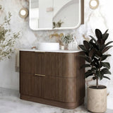 A modern bathroom vanity with a curved wood cabinet, white countertop, integrated white sink, and a large rounded rectangular mirror mounted on a marble wall. A potted plant sits to the right, and the scene is styled as a clean, upscale interior.