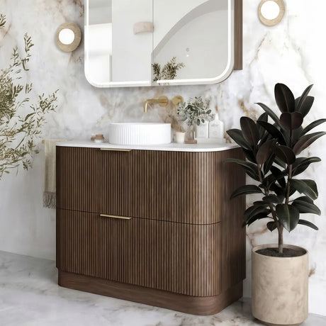 A modern bathroom vanity with a curved wood cabinet, white countertop, integrated white sink, and a large rounded rectangular mirror mounted on a marble wall. A potted plant sits to the right, and the scene is styled as a clean, upscale interior.