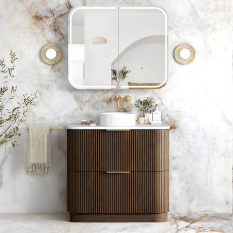A modern bathroom vanity set featuring a ribbed brown wooden cabinet with a white ceramic vessel sink, a white wall-mounted mirror, and a marble-look wall and floor in a bright, styled bathroom setting.