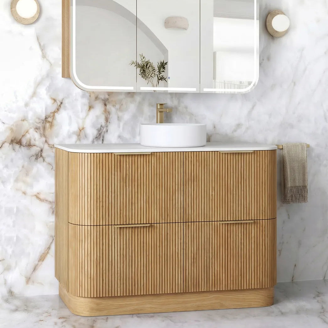 A modern bathroom vanity set featuring a light natural wood ribbed cabinet, a white countertop with a white vessel sink and gold faucet, a three-panel wall-mounted mirror with integrated lighting, and a white marble wall and floor backdrop.