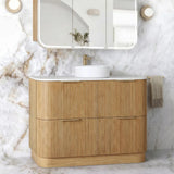 A modern bathroom vanity set featuring a light natural wood ribbed cabinet, a white countertop with a white vessel sink and gold faucet, a three-panel wall-mounted mirror with integrated lighting, and a white marble wall and floor backdrop.