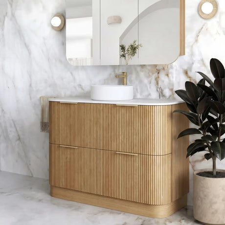 A modern bathroom vanity with a fluted light wood cabinet, white countertop, white vessel sink, and a matching white medicine cabinet mirror mounted on a white marble wall. A potted plant sits to the right.