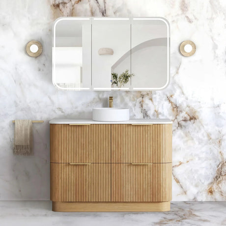 A modern bathroom vanity with a fluted light wood cabinet, white countertop, white round sink, and a large rectangular mirror with a white frame, set against a white marble wall.