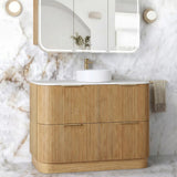 A modern bathroom vanity set featuring a light natural wood ribbed cabinet, a white countertop with a white vessel sink and gold faucet, a three-panel wall-mounted mirror with integrated lighting, and a white marble wall and floor backdrop.