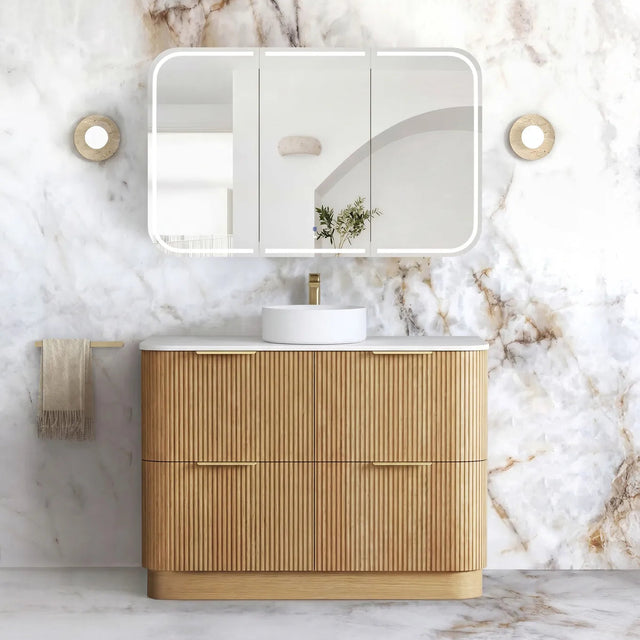 A modern bathroom vanity with a fluted light wood cabinet, white countertop, white round sink, and a large rectangular mirror with a white frame, set against a white marble wall.
