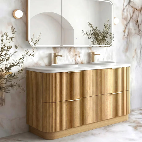 A modern double-sink bathroom vanity with a fluted light wood cabinet, white countertop, and a large three-panel mirror mounted on a marble-look wall. Gold faucets and wall-mounted lights are visible, with decorative branches placed near the mirror and sink area.