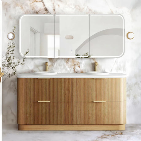 A modern double-sink bathroom vanity with a fluted light wood cabinet, white countertop, and a large three-panel mirror mounted on a white marble wall. Gold-tone faucets and wall-mounted round mirrors are visible, with a marble floor in the foreground.
