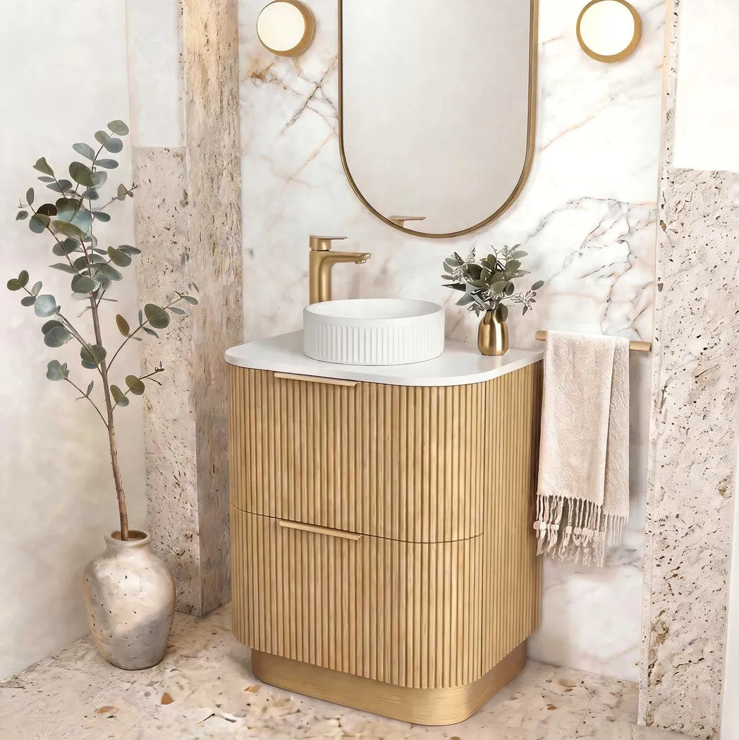 A modern bathroom vanity with a fluted gold cabinet, white marble countertop, and a white vessel sink. An oval gold-framed mirror hangs above on a marble-look wall set within a stone archway, with a gold faucet, small plant, and draped towel nearby.