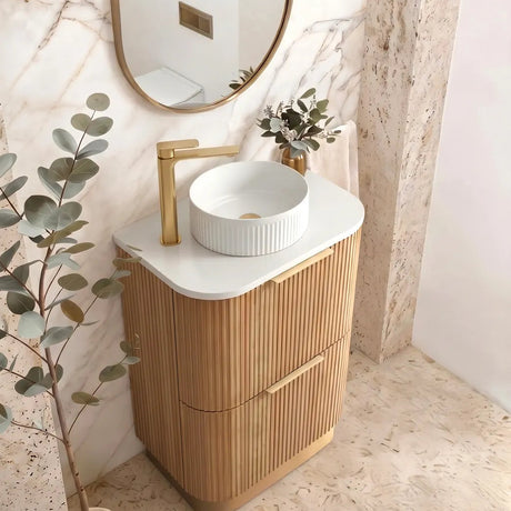 A modern bathroom vanity featuring a fluted light wood cabinet, a white ceramic vessel sink, a gold faucet, and an oval mirror with a gold frame, set against a marble-patterned wall.