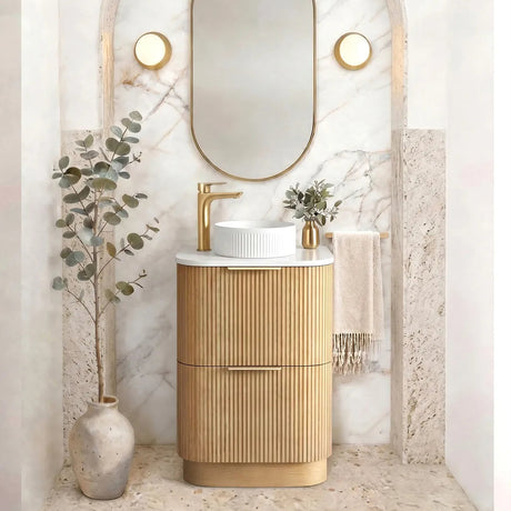A modern bathroom vanity featuring a ribbed gold cabinet, a white vessel sink, a gold faucet, and an arched marble mirror with gold trim, styled with a vase of eucalyptus and a draped towel.