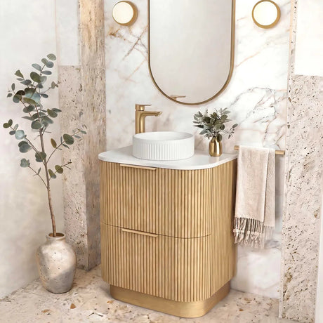 A modern bathroom vanity with a fluted gold cabinet, white marble countertop, and a white vessel sink. An oval gold-framed mirror hangs above on a marble-look wall set within a stone archway, with a gold faucet, small plant, and draped towel nearby.