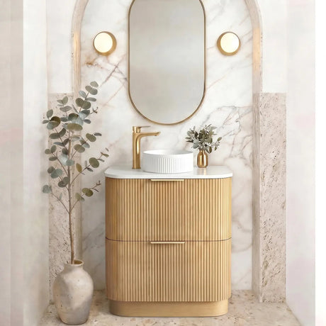 A modern bathroom vanity with a ribbed gold cabinet, white marble top, a white vessel sink, and a matching oval mirror set within an arched marble wall niche.