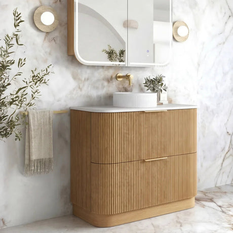 A modern bathroom vanity with a fluted light wood cabinet, white countertop, white vessel sink, and a matching wall-mounted mirror with a white frame, set against a white marble wall.