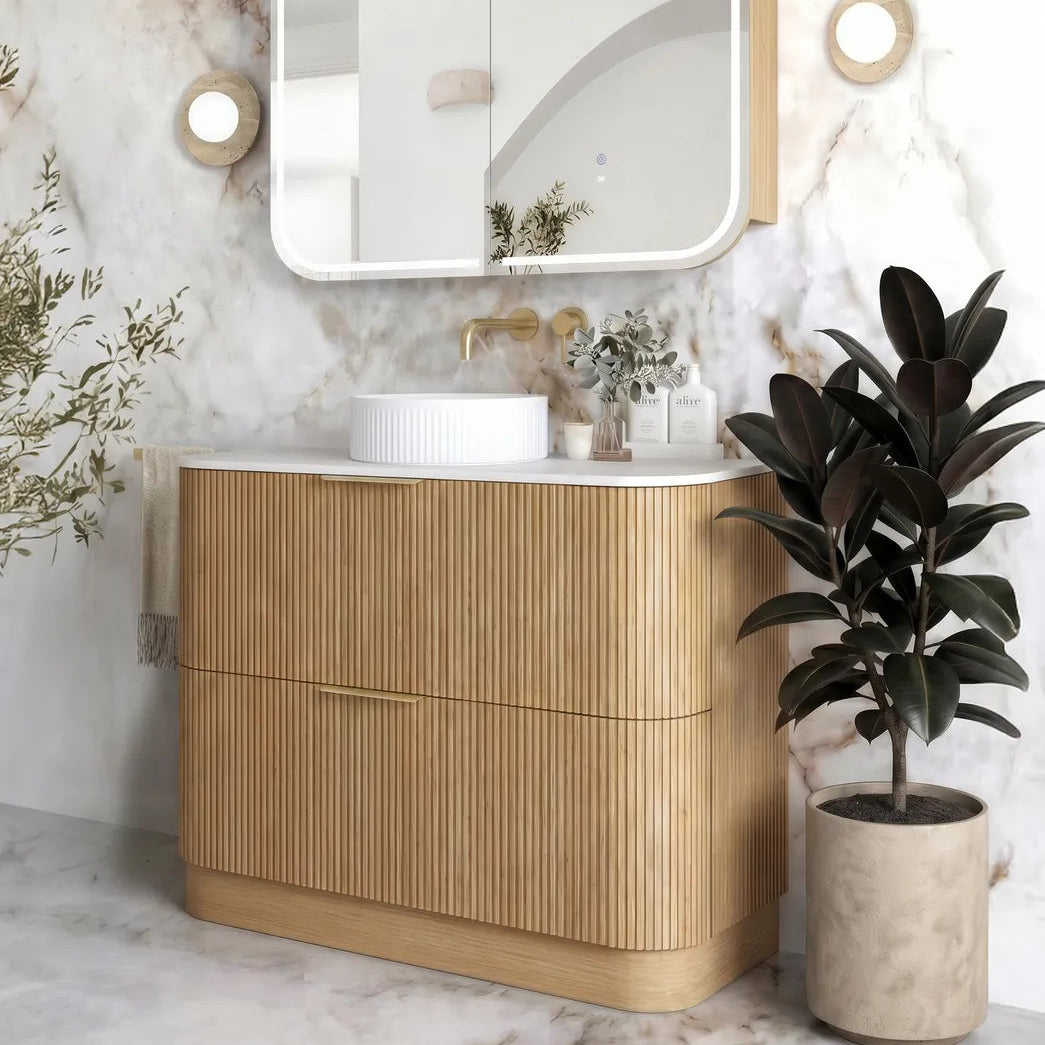 A modern bathroom vanity set featuring a light natural wood cabinet with vertical fluting, a white ceramic vessel sink, a matching wall-mounted mirror with a white frame, and gold-toned fixtures, set against a white marble wall with a potted rubber plant.