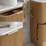 A modern bathroom vanity with fluted light wood cabinets, white marble countertops, a white round vessel sink, and a gold faucet, shown in a clean interior setting with marble walls and a hanging towel.