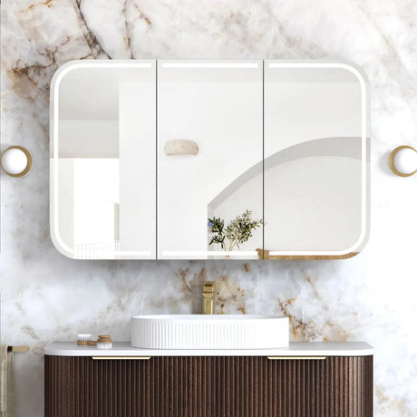 Bathroom vanity with a white sink, wooden cabinet, and three-panel mirror against a marble wall.