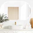 LED Noosa frameless mirror (1200x750) featuring soft ambient backlighting, mounted above a modern white vanity with arched panel details. The minimalist setup includes a round basin, gold faucet, and natural accents like eucalyptus and wicker.