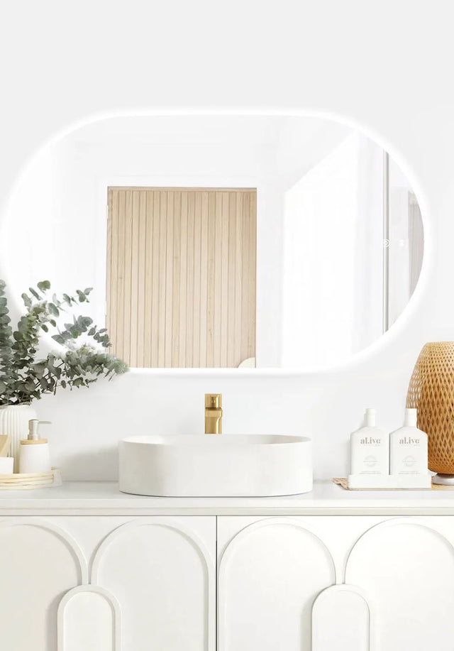 LED Noosa frameless mirror (1200x750) featuring soft ambient backlighting, mounted above a modern white vanity with arched panel details. The minimalist setup includes a round basin, gold faucet, and natural accents like eucalyptus and wicker.