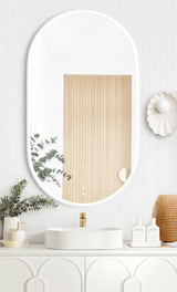 LED Noosa mirror (1200x750) with a sleek white metal frame, mounted above a modern white vanity. The bathroom setup includes a round basin, gold faucet, eucalyptus in a decorative vase, and al.ive hand soap bottles, creating a minimalist and elegant aesthetic.