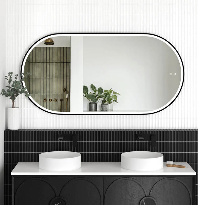 LED Noosa mirror (1500x750) with a sleek black metal frame, mounted above a modern black vanity featuring arched detailing. The bathroom setup includes dual round basins, matte black faucets, and greenery accents, creating a striking monochromatic design.