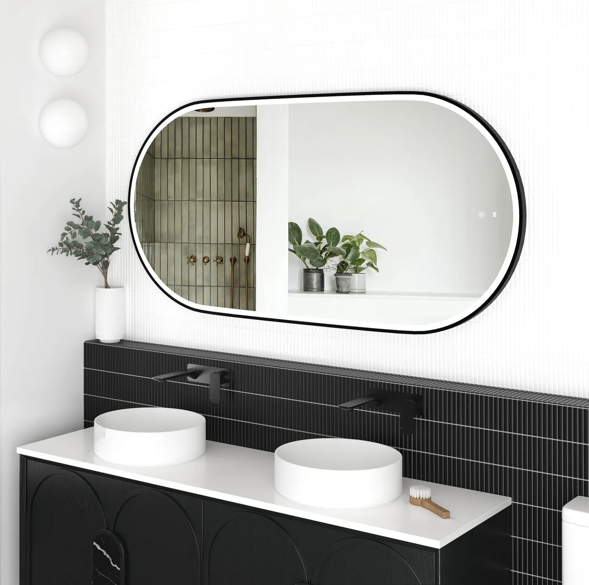 Side view of the LED Noosa 1500x750 black metal frame mirror, highlighting its frameless design and soft backlighting. The setup features a contemporary black vanity with arched paneling, matte black faucets, and decorative plants, exuding minimalist elegance.