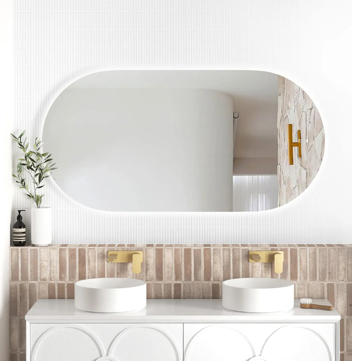 LED Noosa frameless mirror (1500x750) with soft backlighting, mounted above a modern vanity. The bathroom setup features a textured brick backsplash, dual round basins, gold wall-mounted faucets, and minimalist décor including a white vase with greenery.