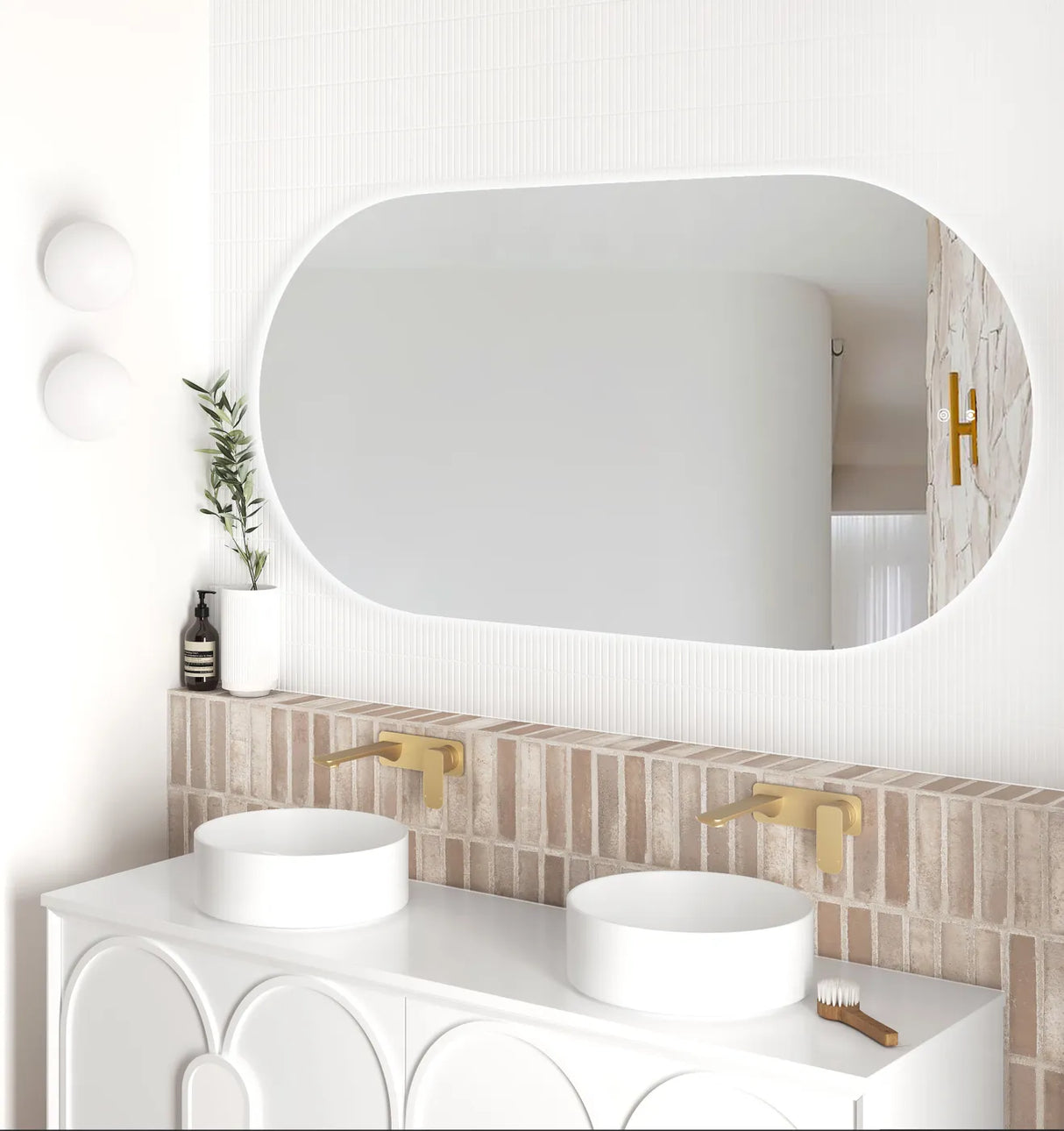 Side angle of the LED Noosa frameless mirror (1500x750), showcasing soft backlighting and a clean, modern bathroom design. The setup includes dual basins, gold wall-mounted faucets, and accent lighting with white globe sconces.