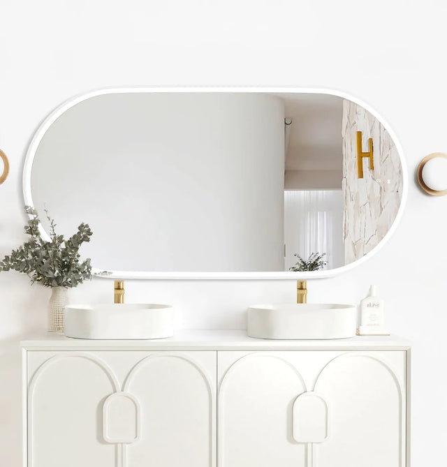 LED Noosa mirror (1500x750) with a white metal frame, featuring soft backlighting, mounted above a modern white vanity with arched panel detailing. The setup includes dual round basins, gold faucets, eucalyptus in a textured vase, and minimalist décor for a clean and contemporary bathroom design.