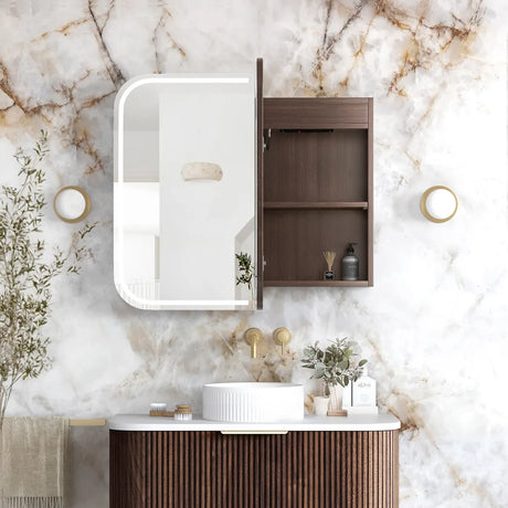 A modern bathroom vanity setup with a dark brown wood cabinet featuring an integrated illuminated mirror and open storage shelf, mounted on a white marble wall above a matching wood vanity with a white vessel sink.