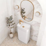 A modern white pedestal sink with a gold faucet, set against a white marble arched wall with a gold-framed oval mirror and wall sconces. A small potted eucalyptus plant sits on the terrazzo-style floor.
